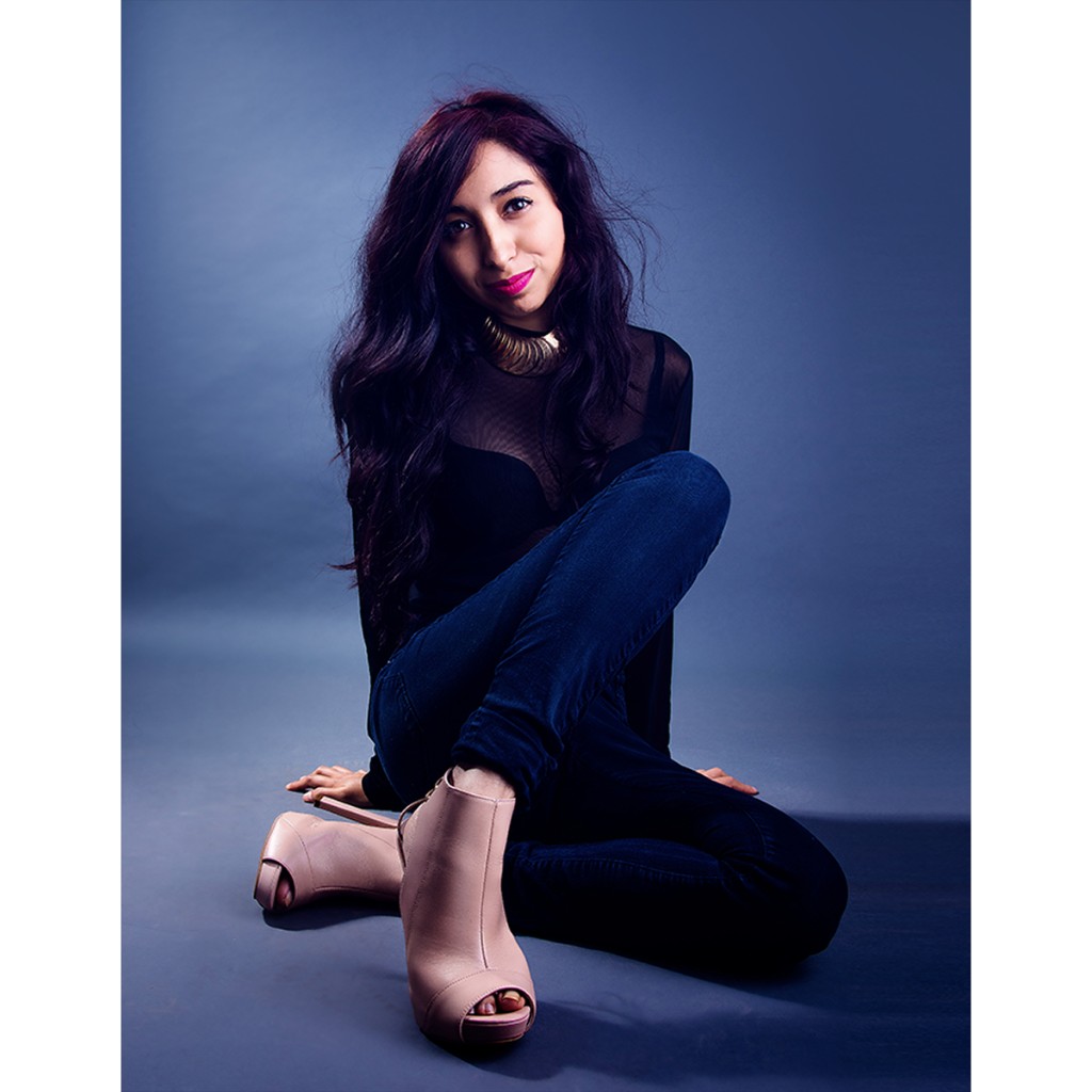 Alondra commercial portrait: seated on the floor with knees raised, sheer black long-sleeve mesh top, dark skinny jeans and nude peep-toe ankle booties; gold disc collar and magenta lip on blue-grey studio backdrop.