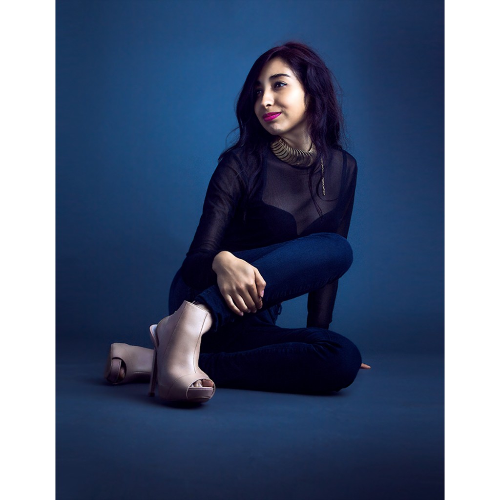 Alondra studio portrait: seated cross-legged in black mesh top and dark jeans, nude peep-toe heels and gold collar; gaze away from camera on saturated blue backdrop — commercial talent session.