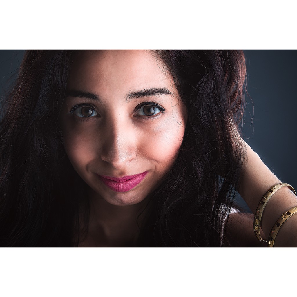 Alondra beauty close-up: face filling the frame, bold magenta lipstick and gold star-engraved bangles; soft studio light on dark charcoal background — commercial talent portrait.