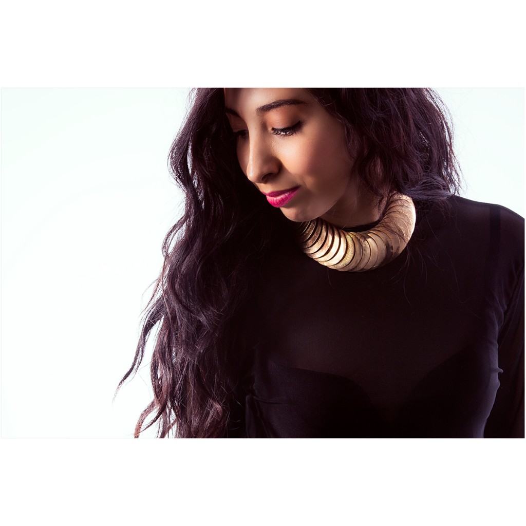 Alondra editorial portrait: head bowed with lowered gaze, black mesh top and large gold disc collar necklace; backlit hair on high-key white backdrop — commercial portrait photography.