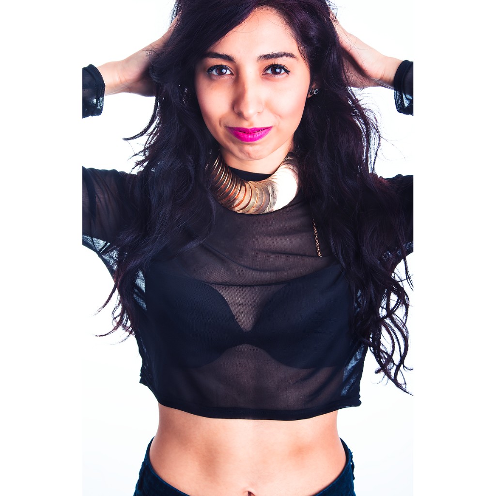 Alondra waist-up portrait: both hands in hair, black sheer mesh crop top over visible bra, gold disc choker and bare midriff; white high-key background — commercial fashion headshot.