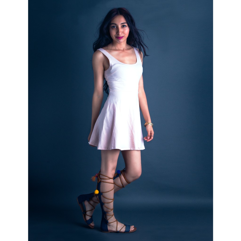 Alondra full-length portrait: white fit-and-flare scoop-neck dress, blue lace-up gladiator sandals with colorful pompoms, gold bangles; windblown hair on deep teal-blue backdrop — talent portrait session.