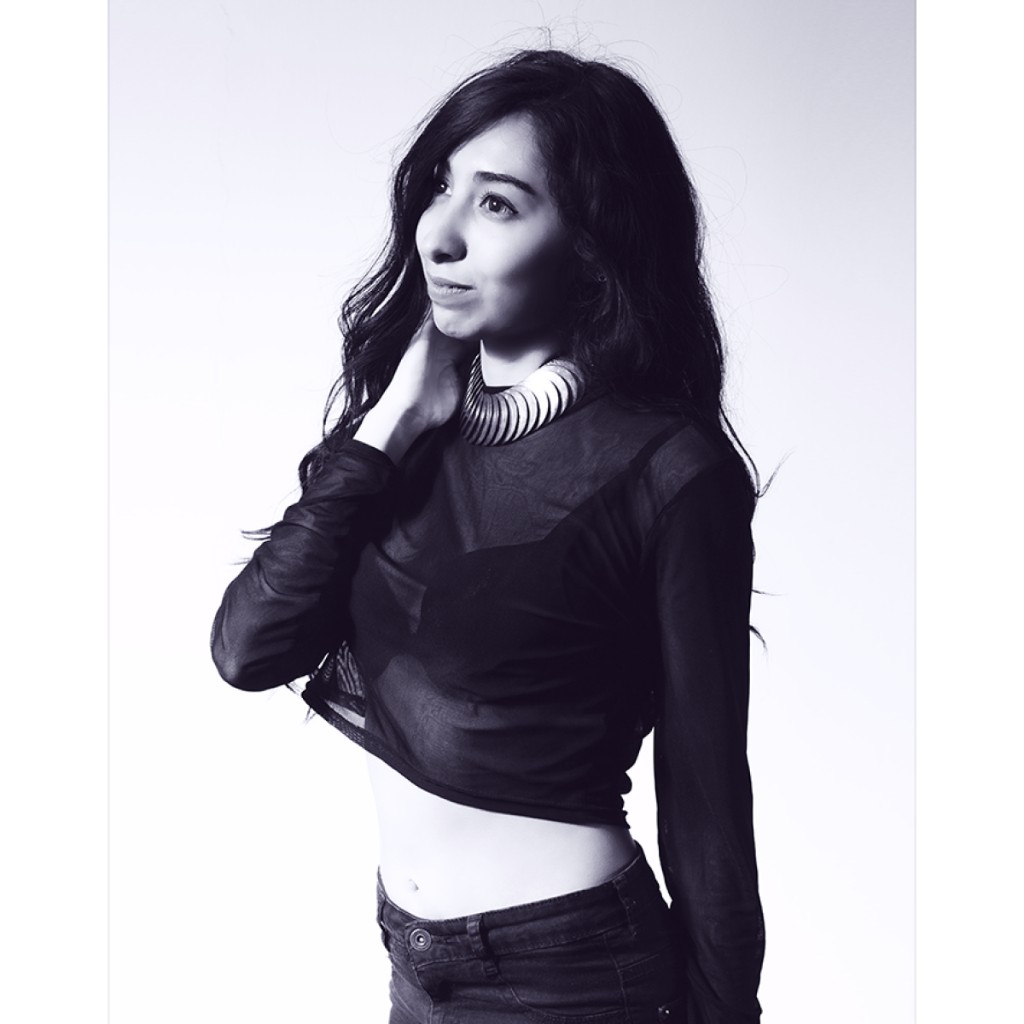 Alondra B&W portrait: black long-sleeve mesh crop top, striped silver band collar and hand at neck; three-quarter profile looking upward on white background — editorial studio photography.