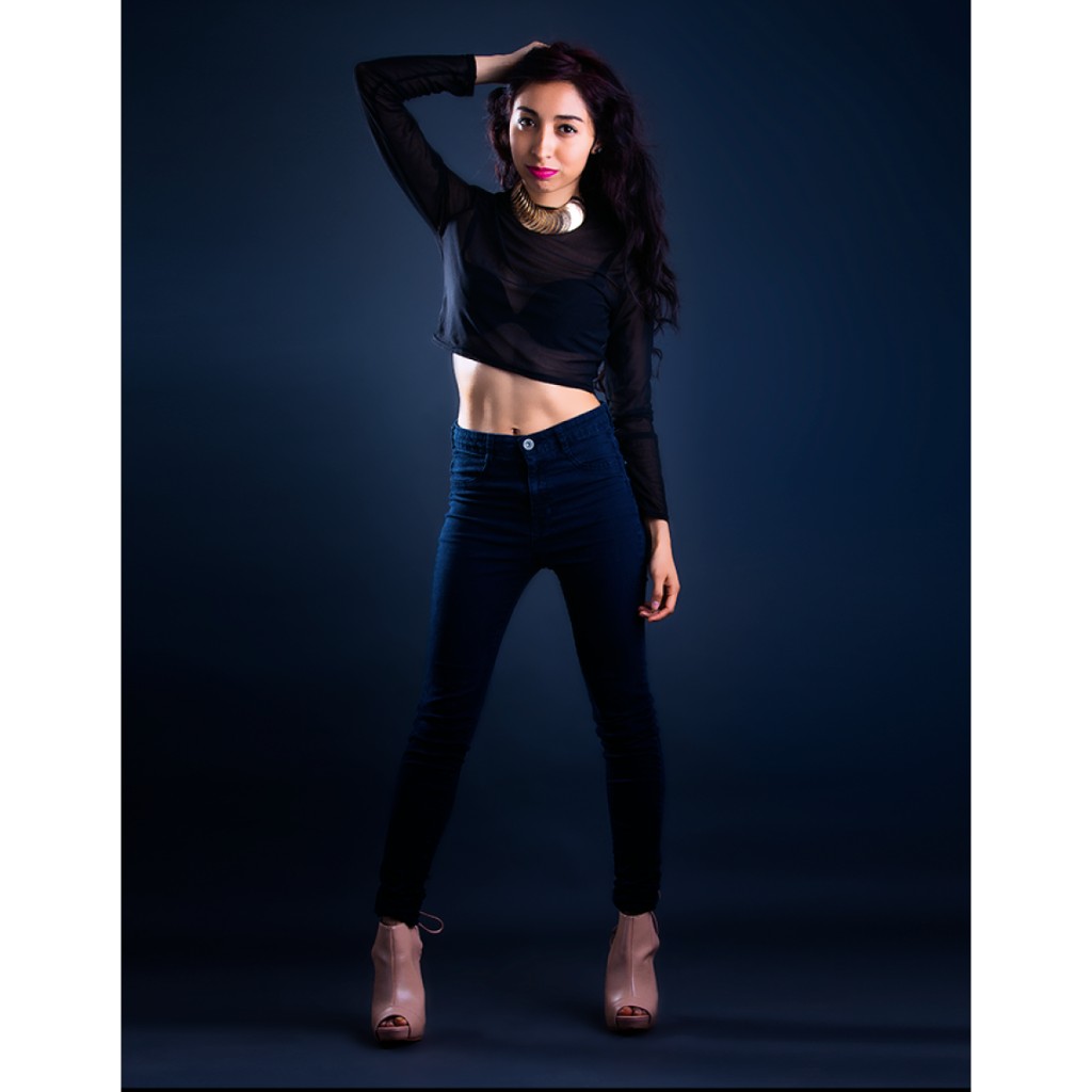 Alondra editorial portrait: black sheer mesh crop top, high-waist dark skinny jeans and nude peep-toe booties, gold disc collar and hand in hair; full-length with visible midriff on navy gradient backdrop.