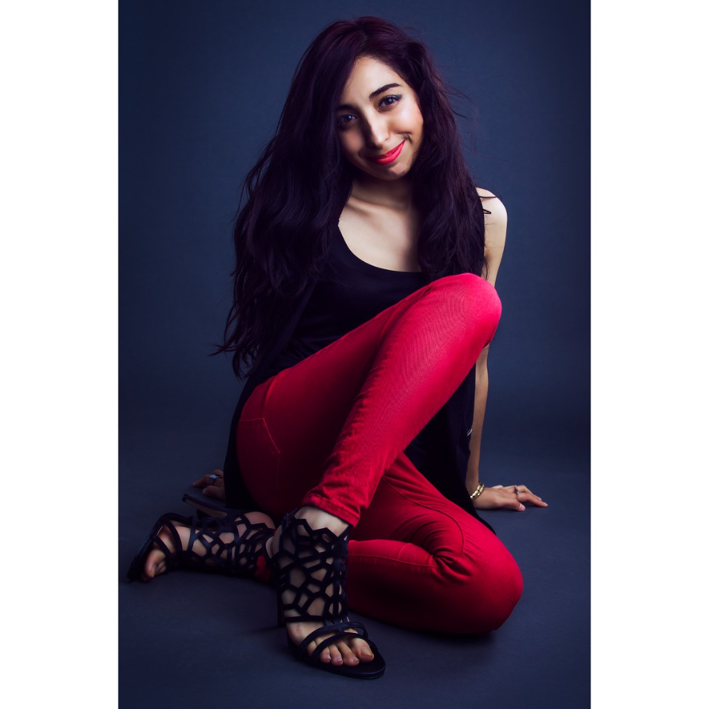 Alondra dynamic portrait: seated on floor with knees up, black sleeveless tank, red skinny jeans and black geometric cut-out sandals; wide smile and gold bangle on blue-grey studio backdrop — commercial portrait session.