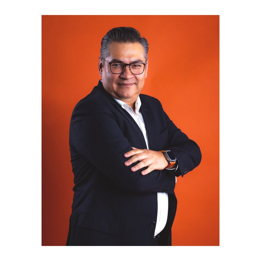Doto corporate portrait: man in navy blazer and white shirt, arms crossed and orange-band watch, orange studio backdrop.