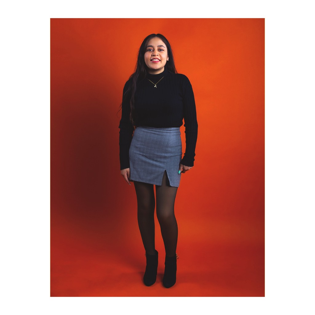 Doto full-length portrait: smiling woman in black turtleneck, grey plaid mini skirt, black tights and boots, orange backdrop.