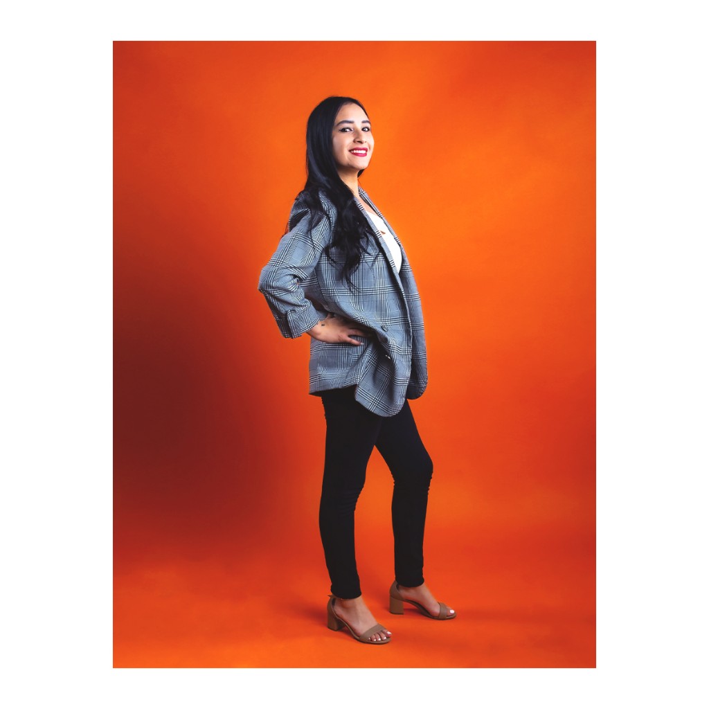 Doto portrait: woman in grey plaid blazer, white top and black pants, tan heeled sandals, orange backdrop.