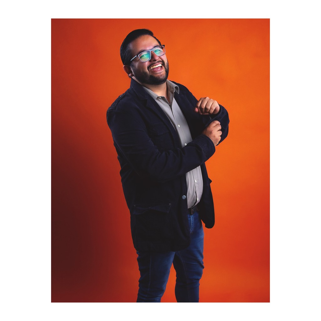 Candid doto portrait: bearded man with glasses laughing, dark blazer, grey shirt and jeans, orange background.
