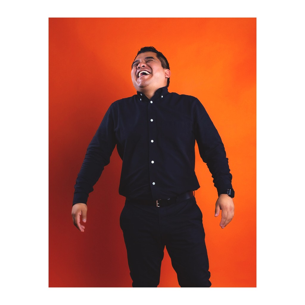Doto lifestyle portrait: man laughing in dark button-down and dark trousers, vibrant orange studio backdrop.