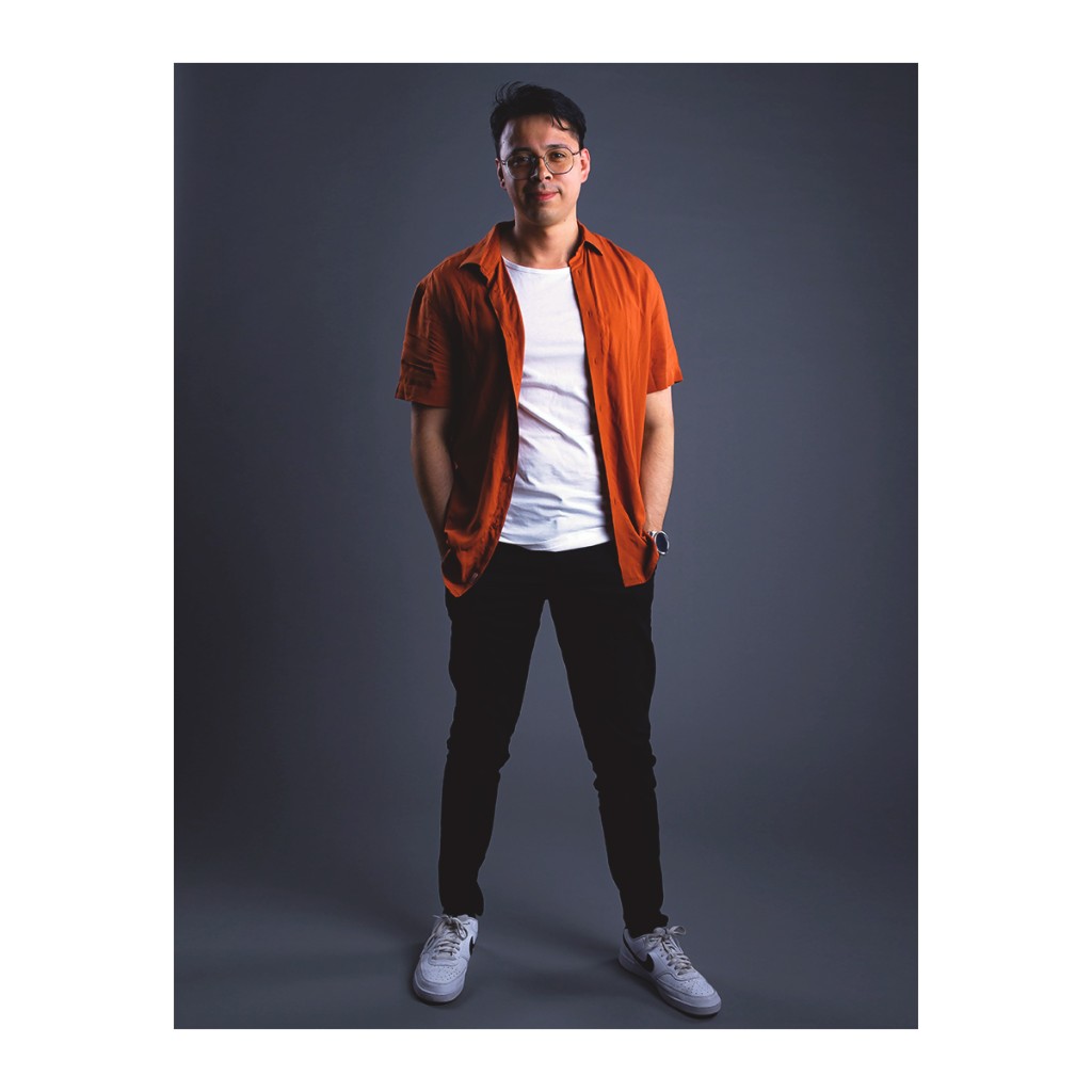 Doto style portrait: young man in open orange shirt over white tee, black pants and white sneakers, grey backdrop.
