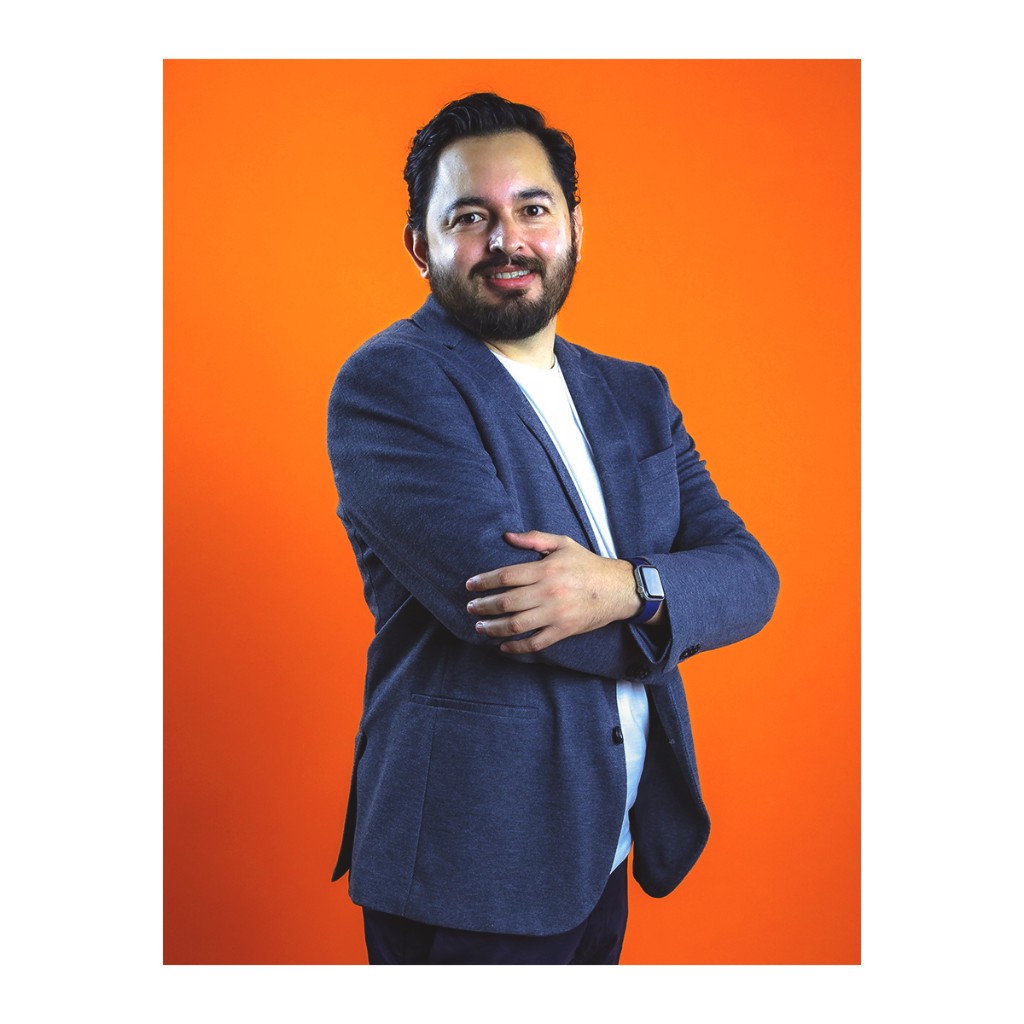 Doto corporate portrait: bearded man in textured blue blazer and white t-shirt, arms crossed, orange background.