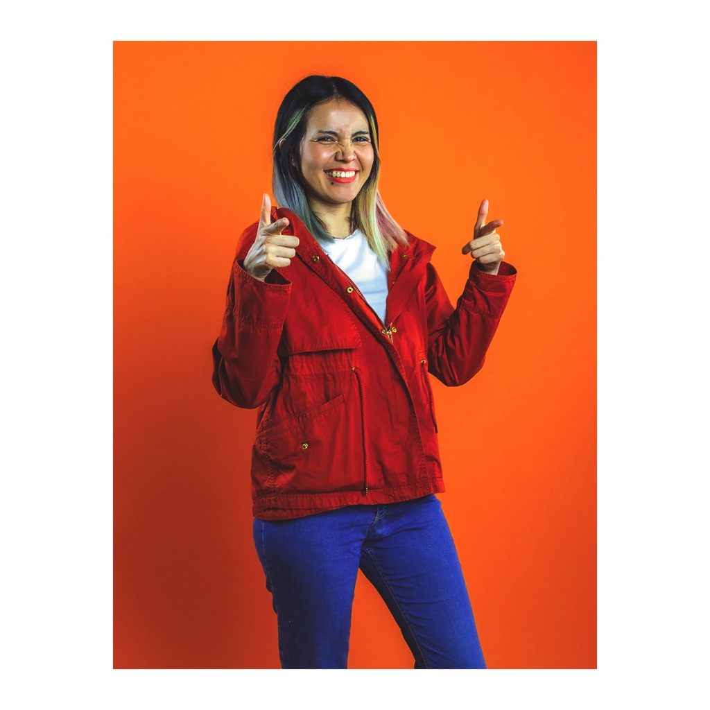 Dynamic doto portrait: woman in red jacket and jeans, blue-tipped hair, playful finger-pointing gesture, orange backdrop.