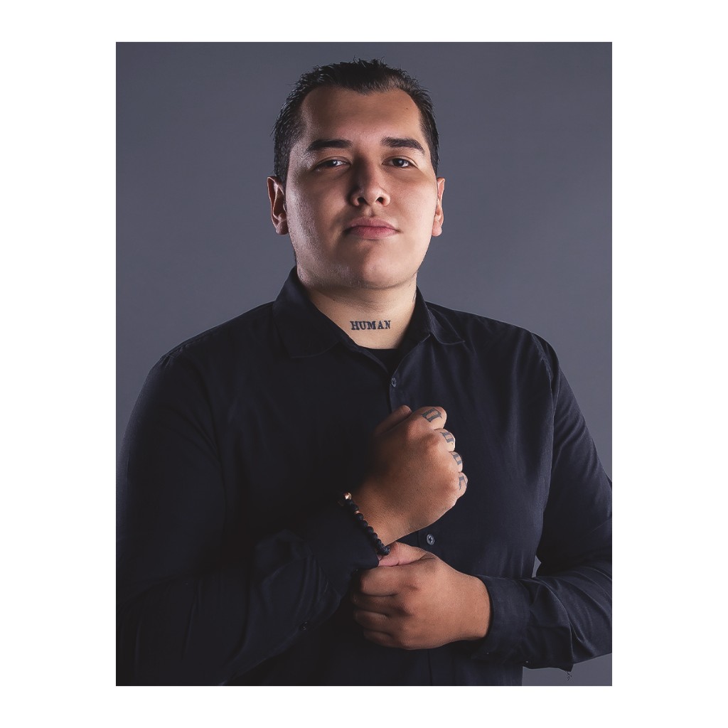 Editorial doto portrait: man in black shirt with HUMAN neck tattoo, fist pose and rim light, dark grey background.