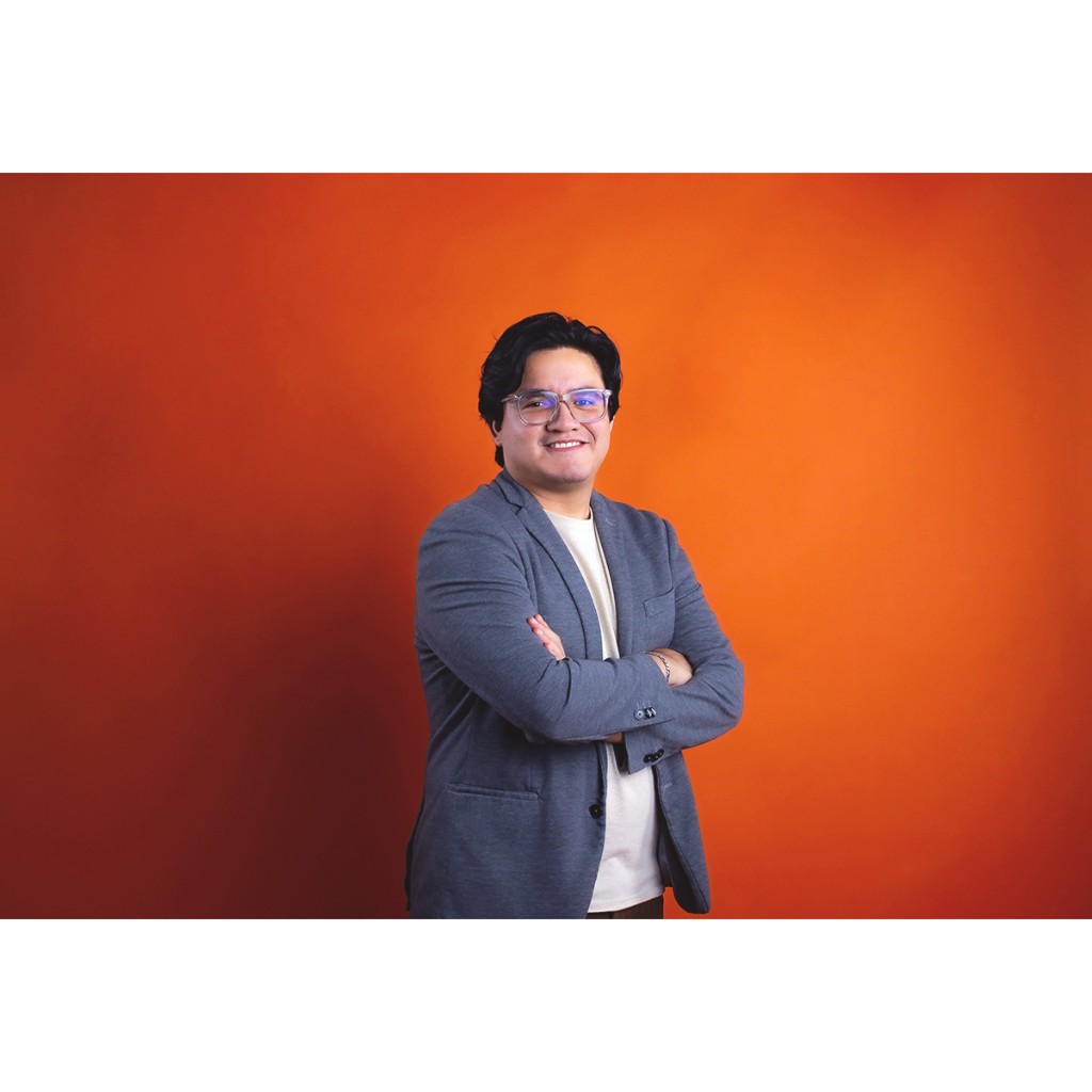Doto portrait: man in clear-frame glasses and grey blazer, smiling with arms crossed, orange background.