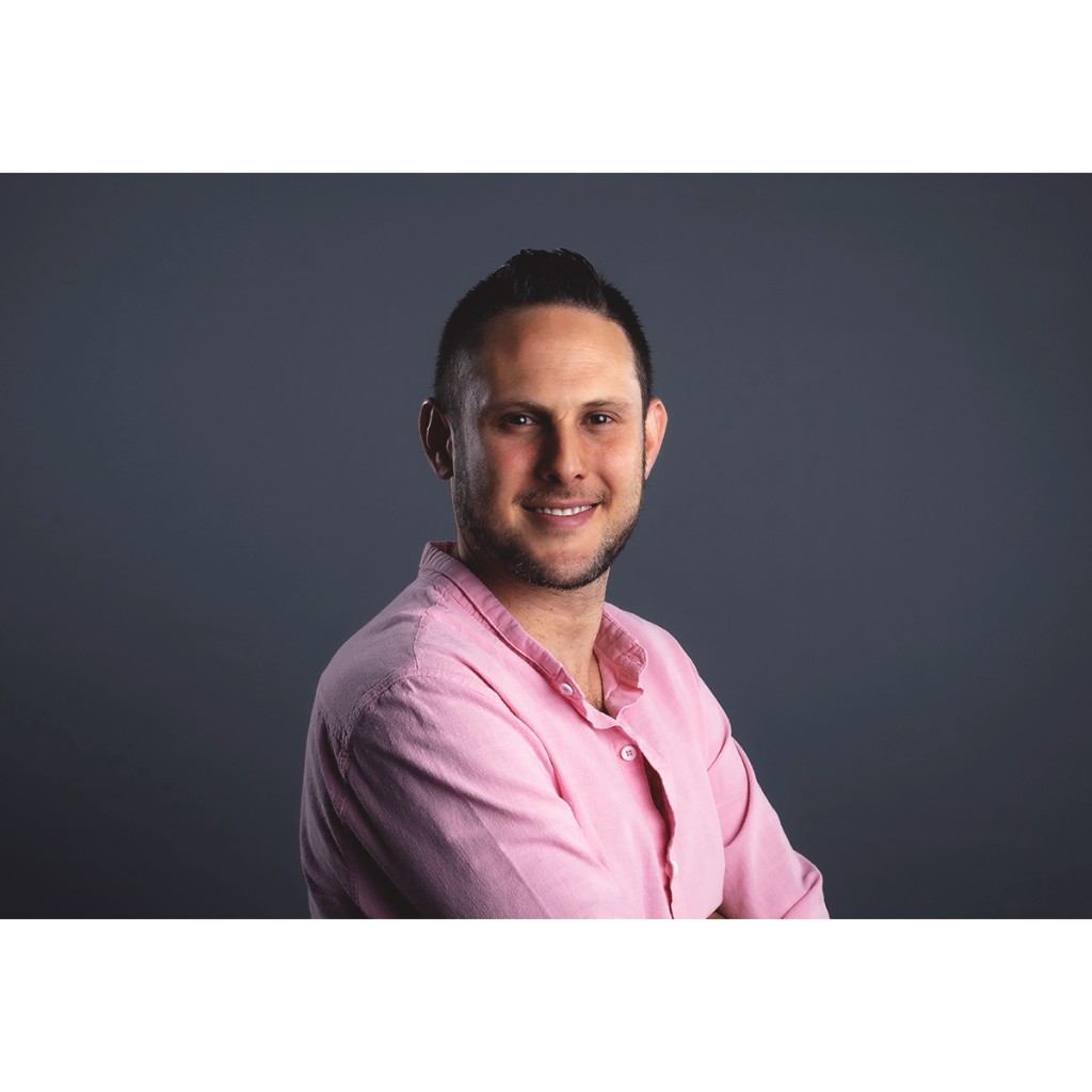 Doto portrait: bearded man in pink button-down, arms crossed, warm expression, dark grey backdrop.