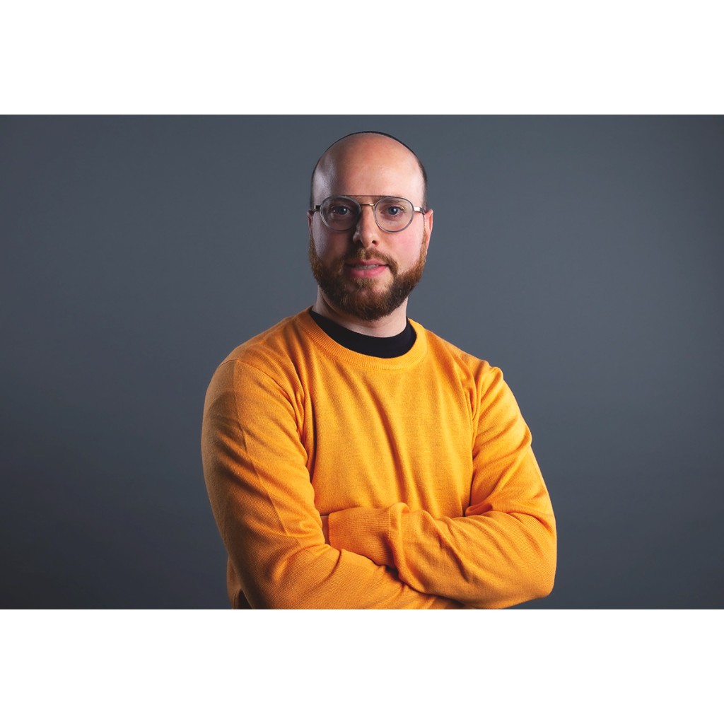 Doto portrait: man in mustard sweater and round glasses, arms crossed, dark grey studio backdrop.