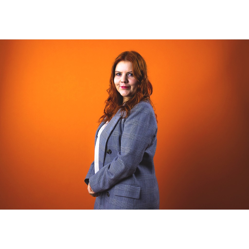Doto portrait: red-haired woman in grey plaid blazer, clasped hands, eye contact, orange background.