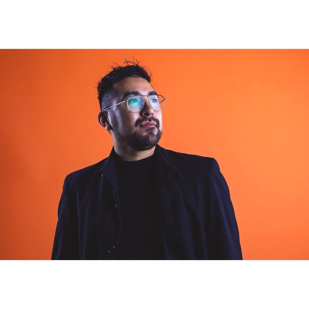 Doto portrait: bearded man in black blazer and turtleneck, metal glasses with lens catchlight, orange backdrop.