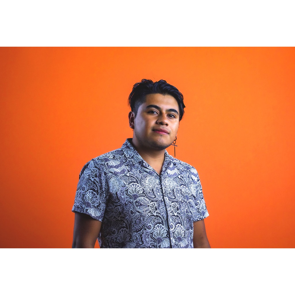 Doto portrait: young man in short-sleeve paisley shirt and cross earring, orange studio background.