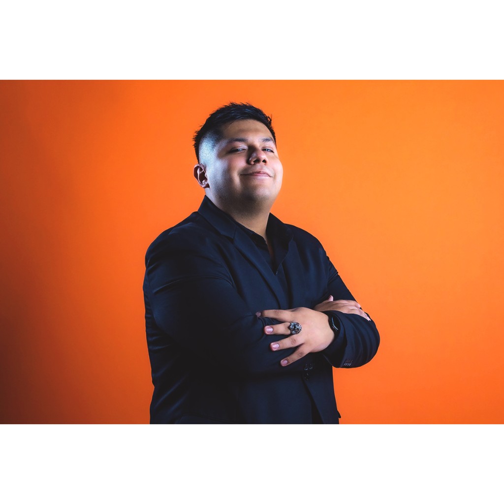 Doto portrait: man in black suit and black shirt, arms crossed, ring and watch, orange background.