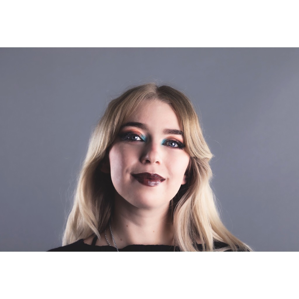 Doto beauty close-up: blonde woman with teal-and-copper artistic eye makeup and chocolate lips, grey backdrop.
