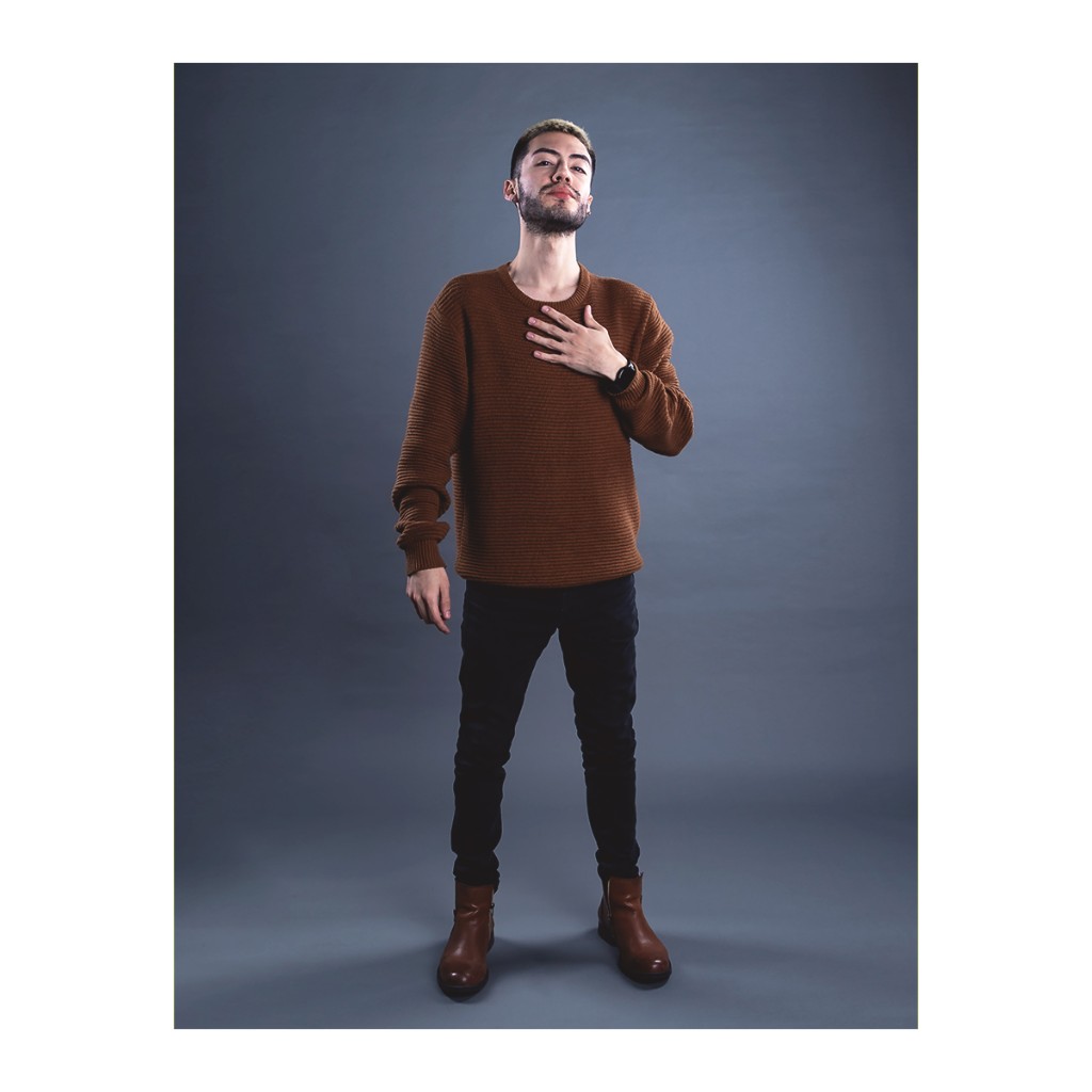 Doto full-length portrait: bearded man in brown knit sweater, black pants and brown Chelsea boots, hand on chest, grey backdrop.