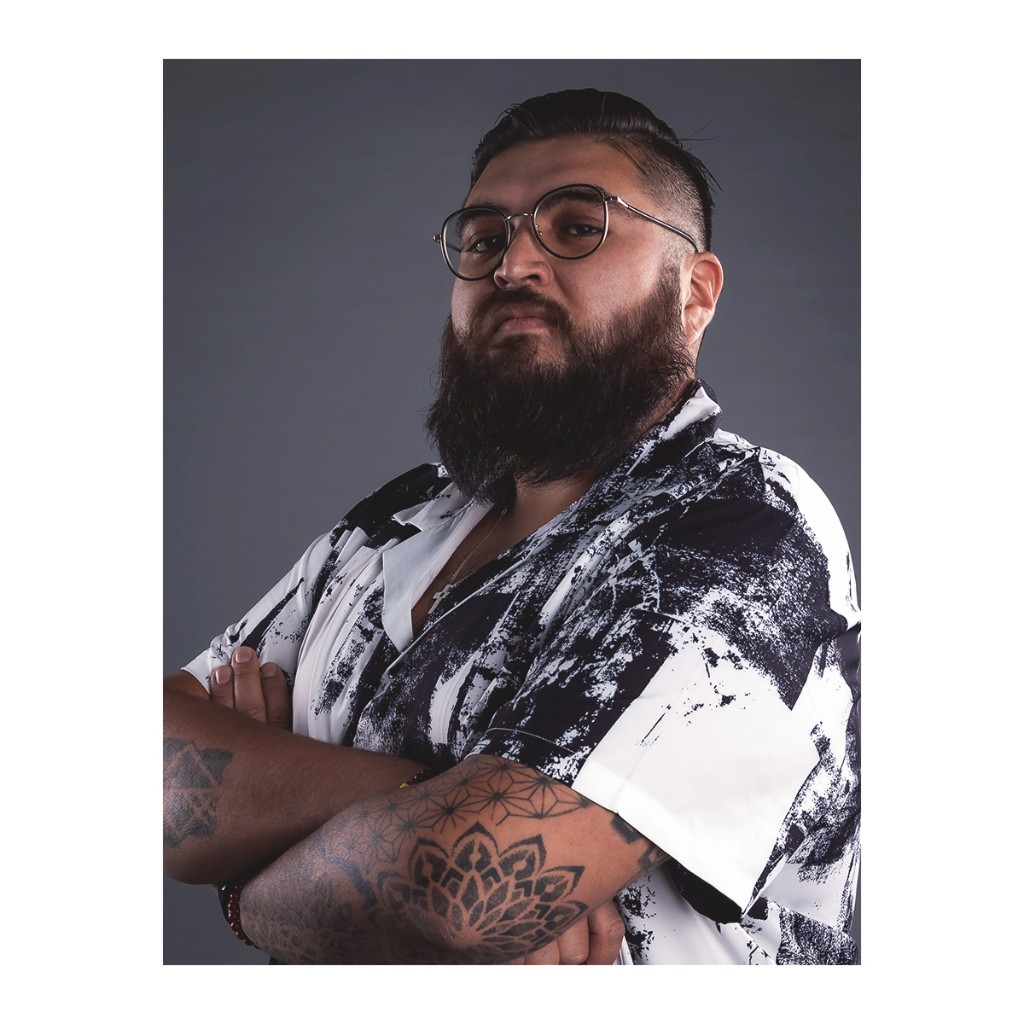 Doto portrait: bearded man in round glasses, black-and-white abstract short-sleeve shirt and mandala forearm tattoos, grey background.