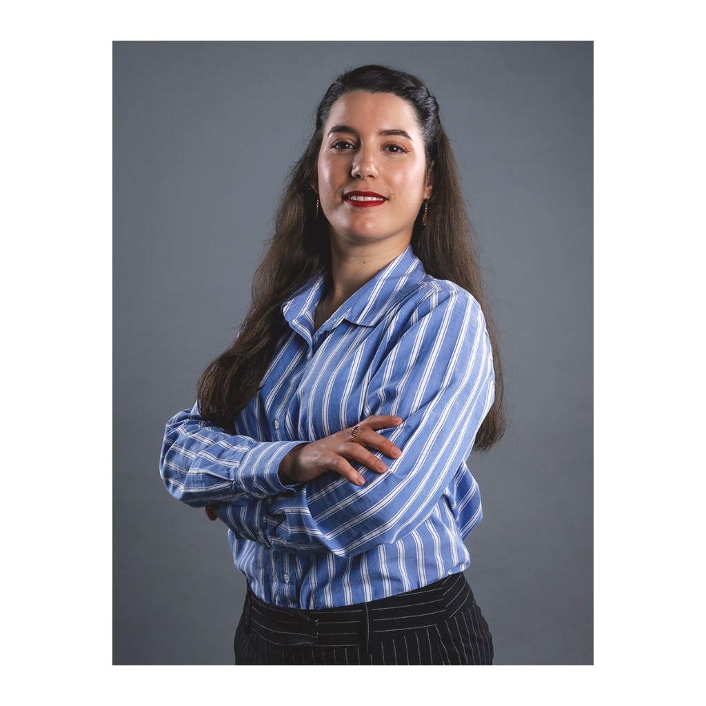 Doto corporate portrait: woman in blue-and-white striped shirt and pinstripe trousers, arms crossed, grey backdrop.