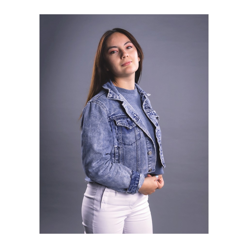 Doto lifestyle portrait: woman in light denim jacket, grey sweater and white pants, grey studio backdrop.