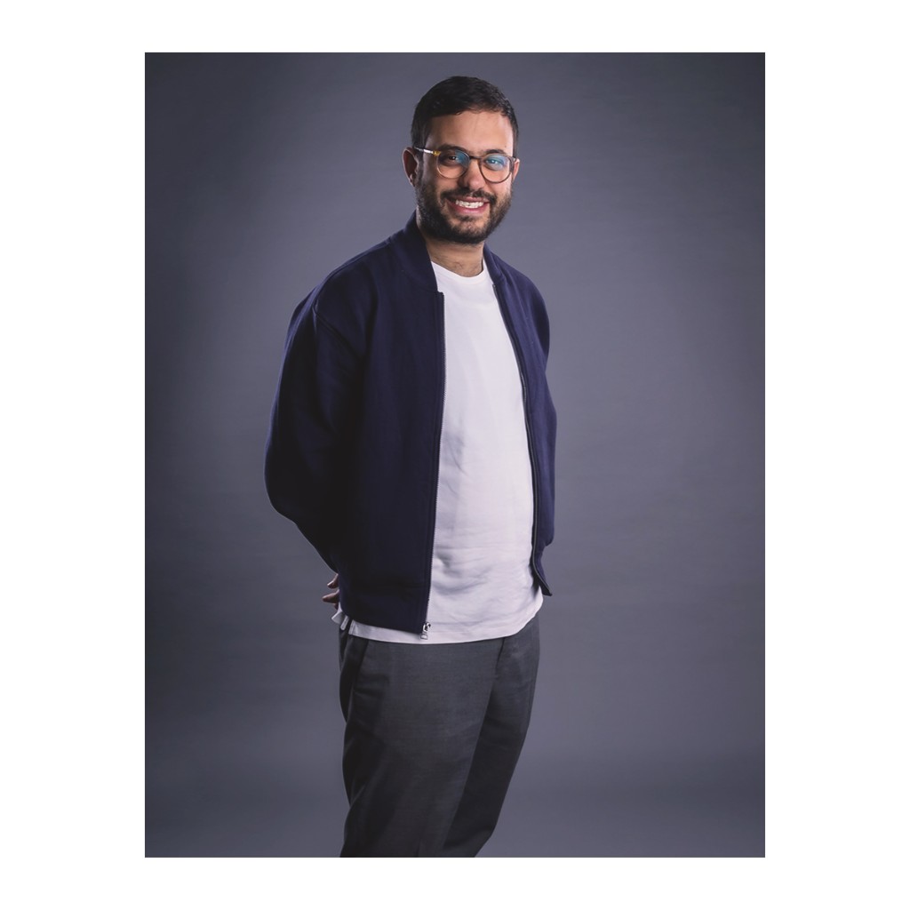 Doto portrait: smiling bearded man in navy bomber over white tee and grey trousers, hands behind back, grey backdrop.
