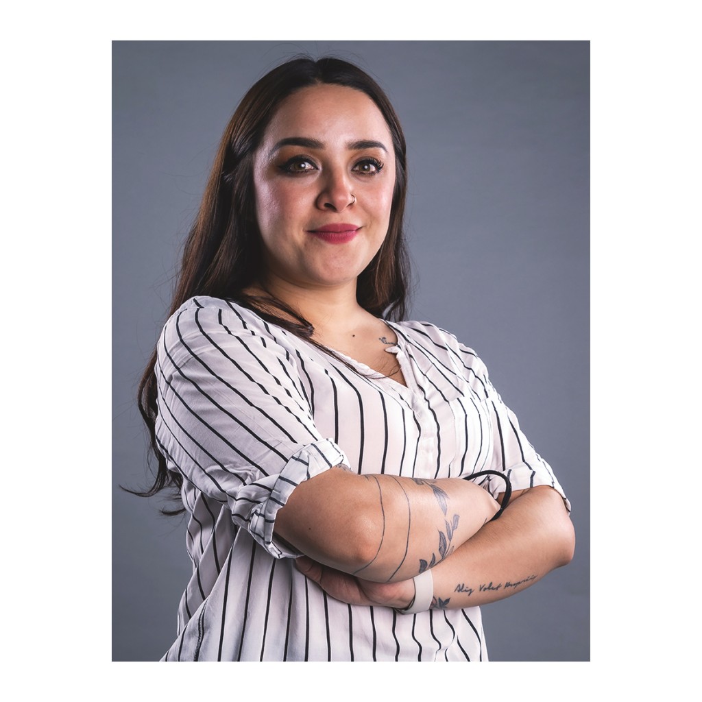 Doto portrait: woman in white pinstripe shirt, forearm tattoos and nose stud, arms crossed, grey background.