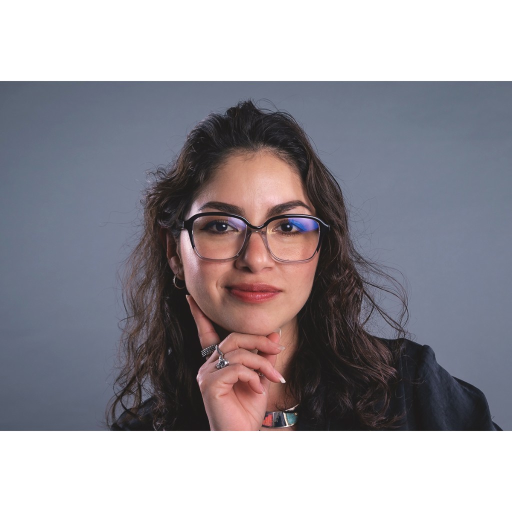 Doto close-up: woman with wavy dark hair, black-framed glasses with blue lens reflection, hand under chin, grey backdrop.