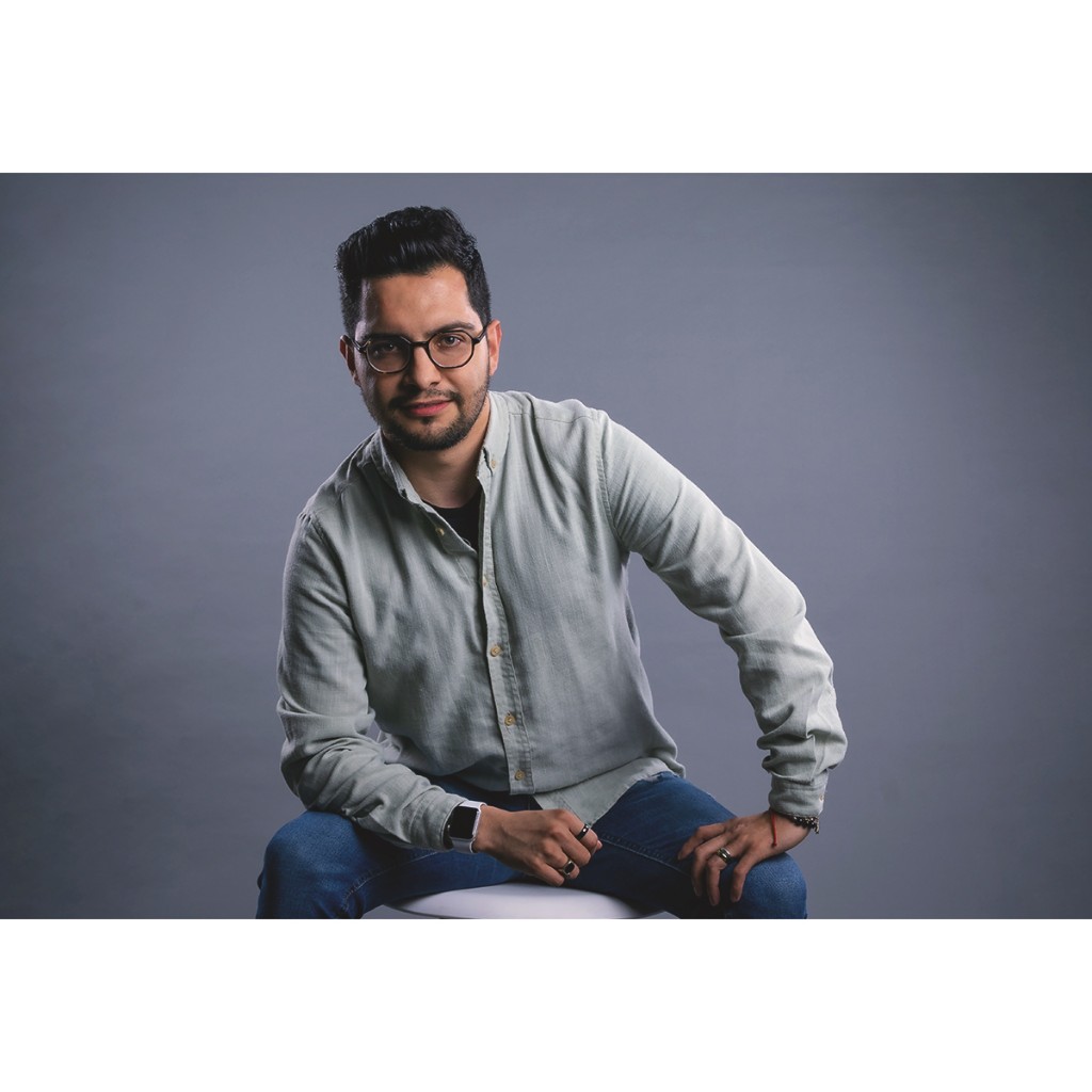 Doto portrait: bearded man on white stool in textured sage shirt and blue jeans, round glasses, grey backdrop.