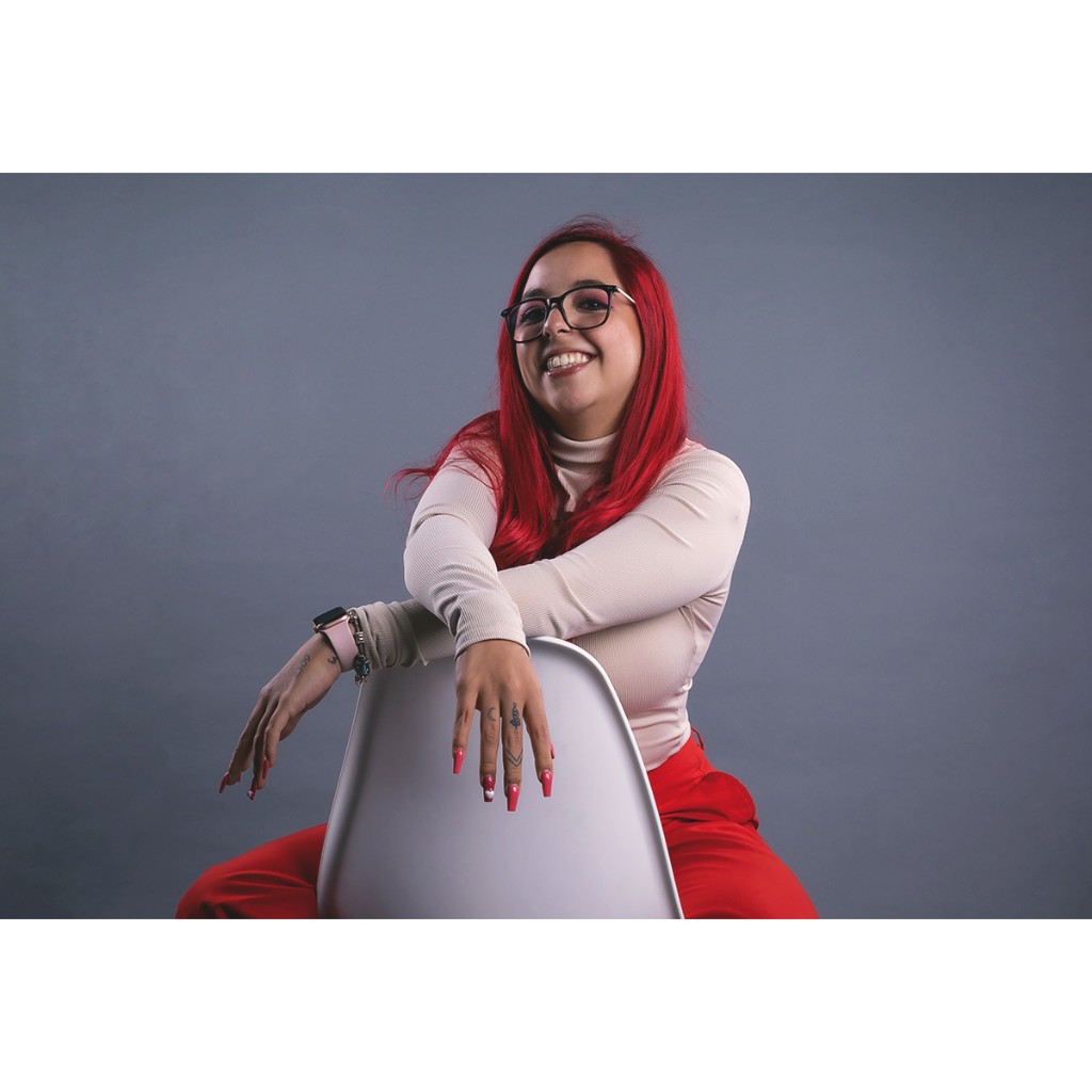 Creative doto portrait: woman with vibrant red hair, black glasses, beige turtleneck and red pants, seated backward on white chair, grey backdrop.