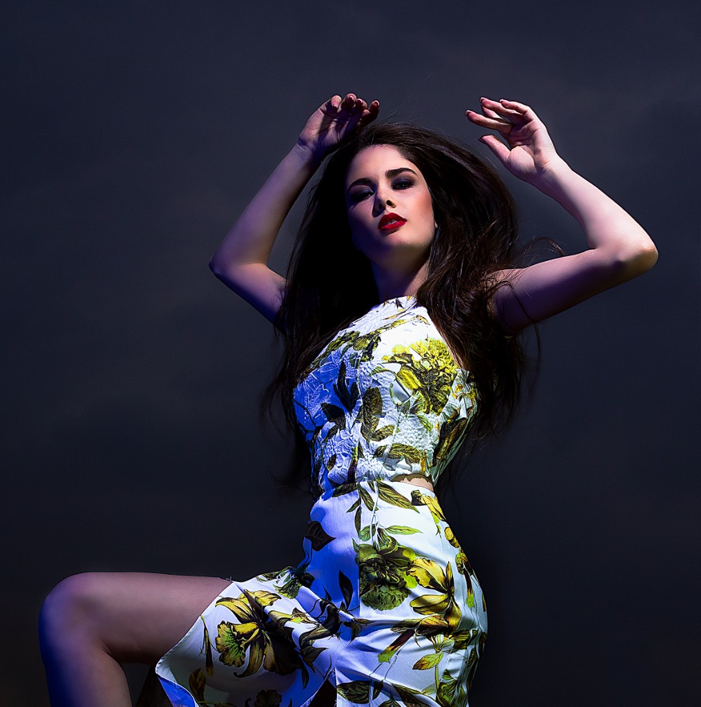 Ferezing editorial portrait: model in white yellow-and-green floral dress, arms raised, blue-violet rim light on dark studio backdrop.