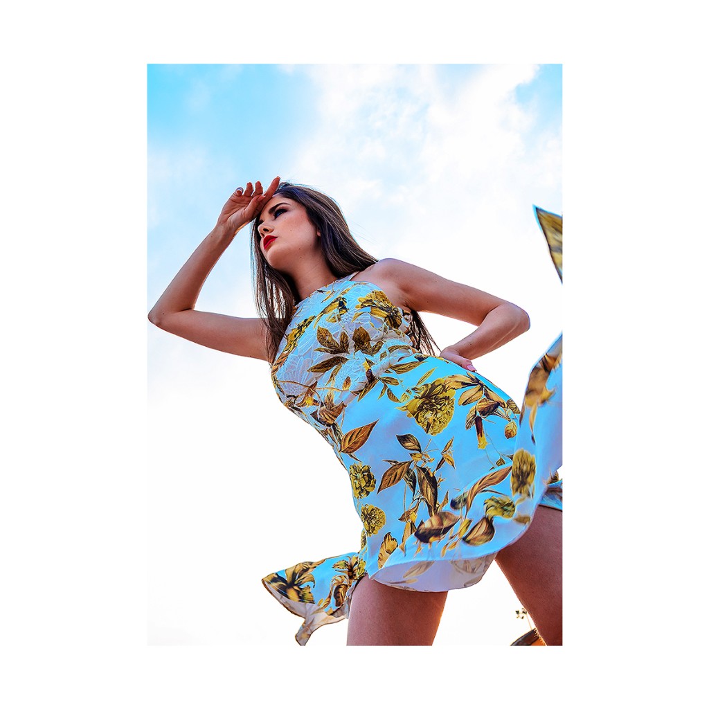 Ferezing outdoor fashion: short halter floral dress, hand at forehead, skirt catching wind, low-angle bright blue sky.