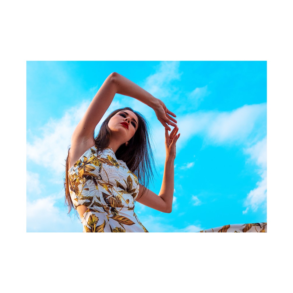 Ferezing low-angle portrait: high-neck white floral dress, arms arched overhead, clear sky with soft clouds.