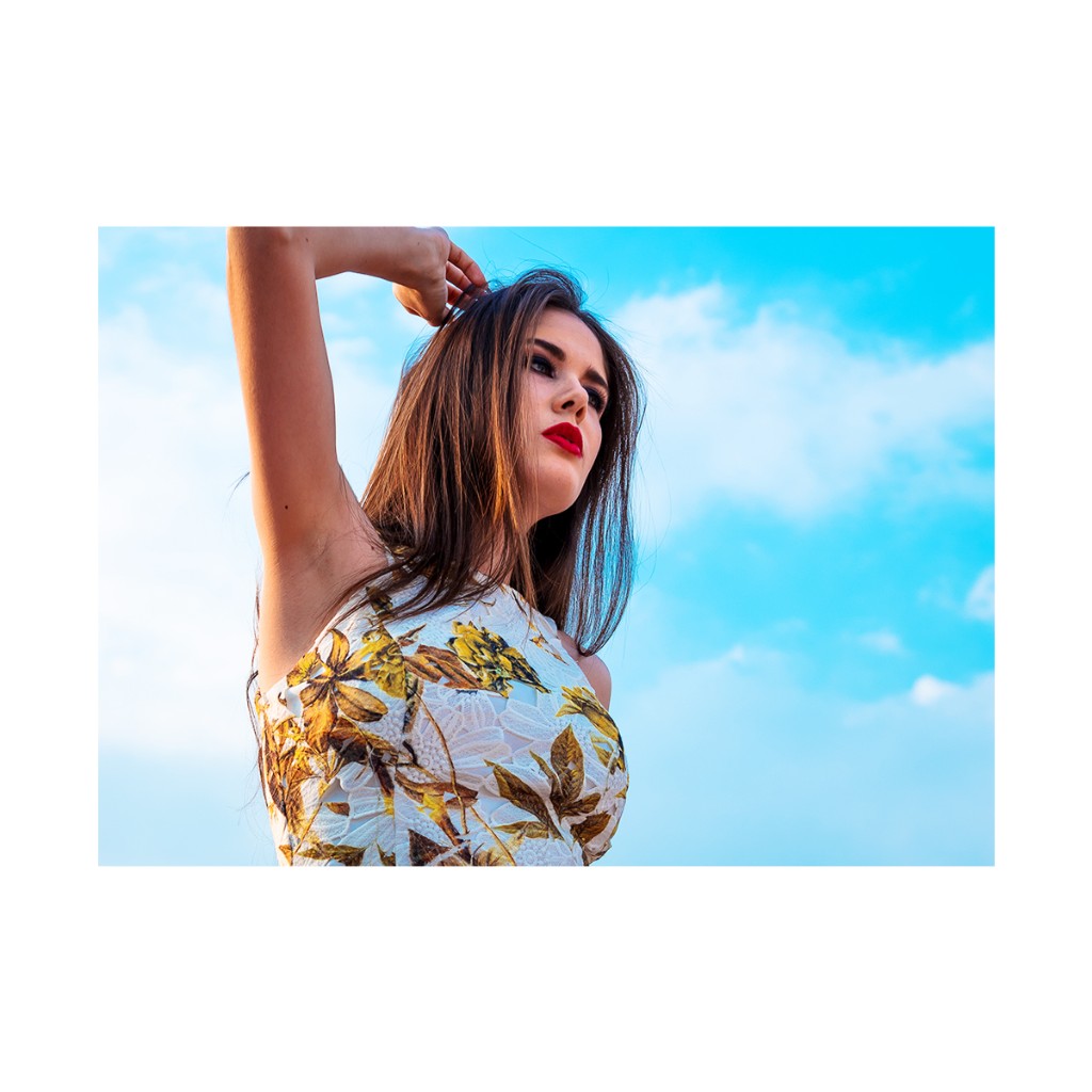 Ferezing portrait: lace-like floral sleeveless top, hand behind head, gaze to the side, blue sky and wispy clouds.