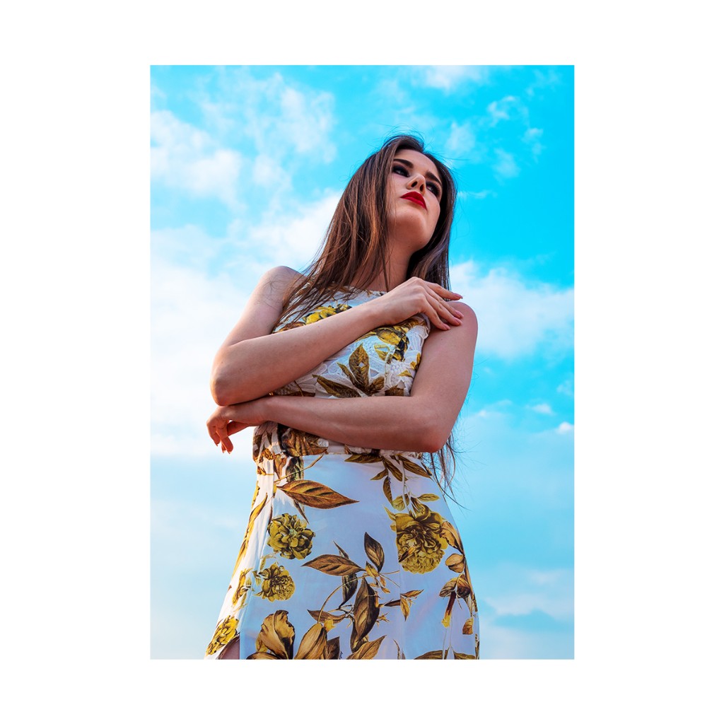 Ferezing portrait: summer floral dress, arms crossed at chest, low angle against saturated blue sky.