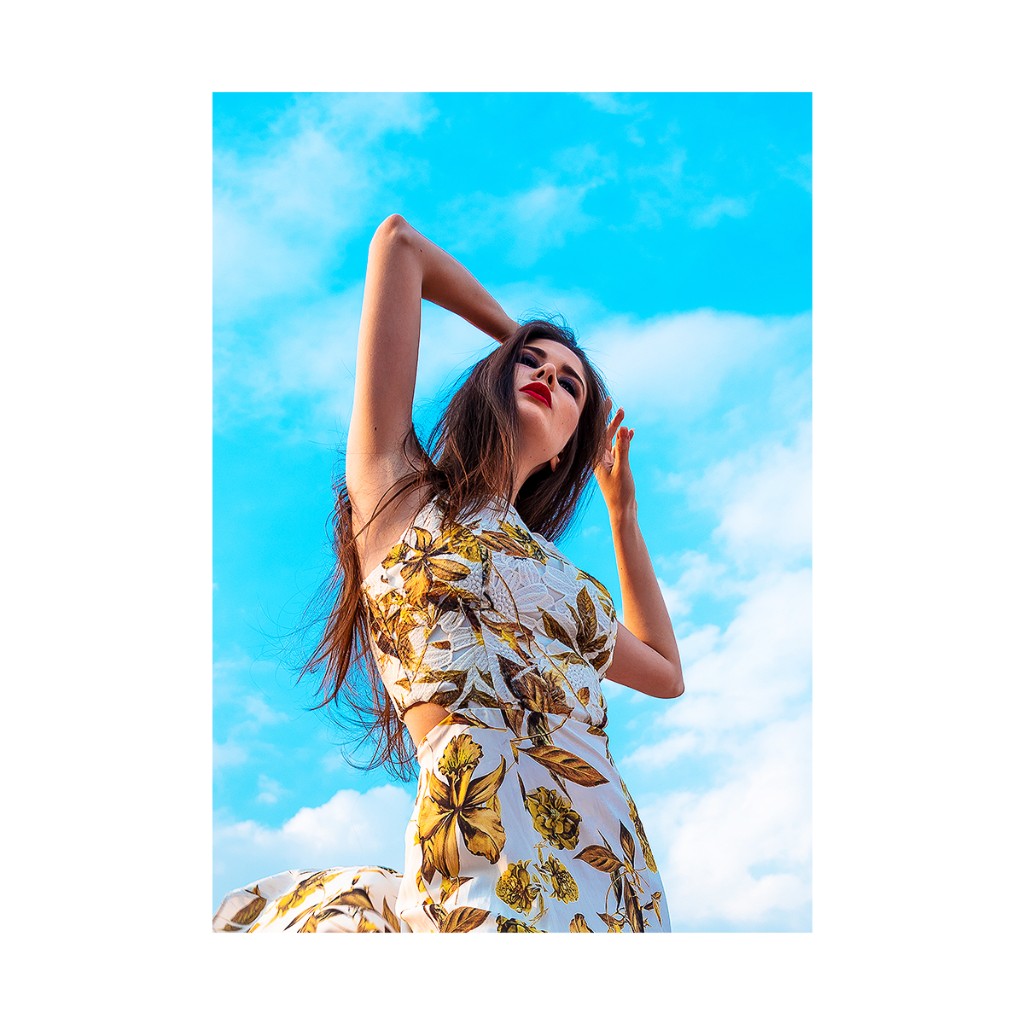 Ferezing fashion: white floral dress with waist cutout, hand on crown, eye contact, vivid blue sky.