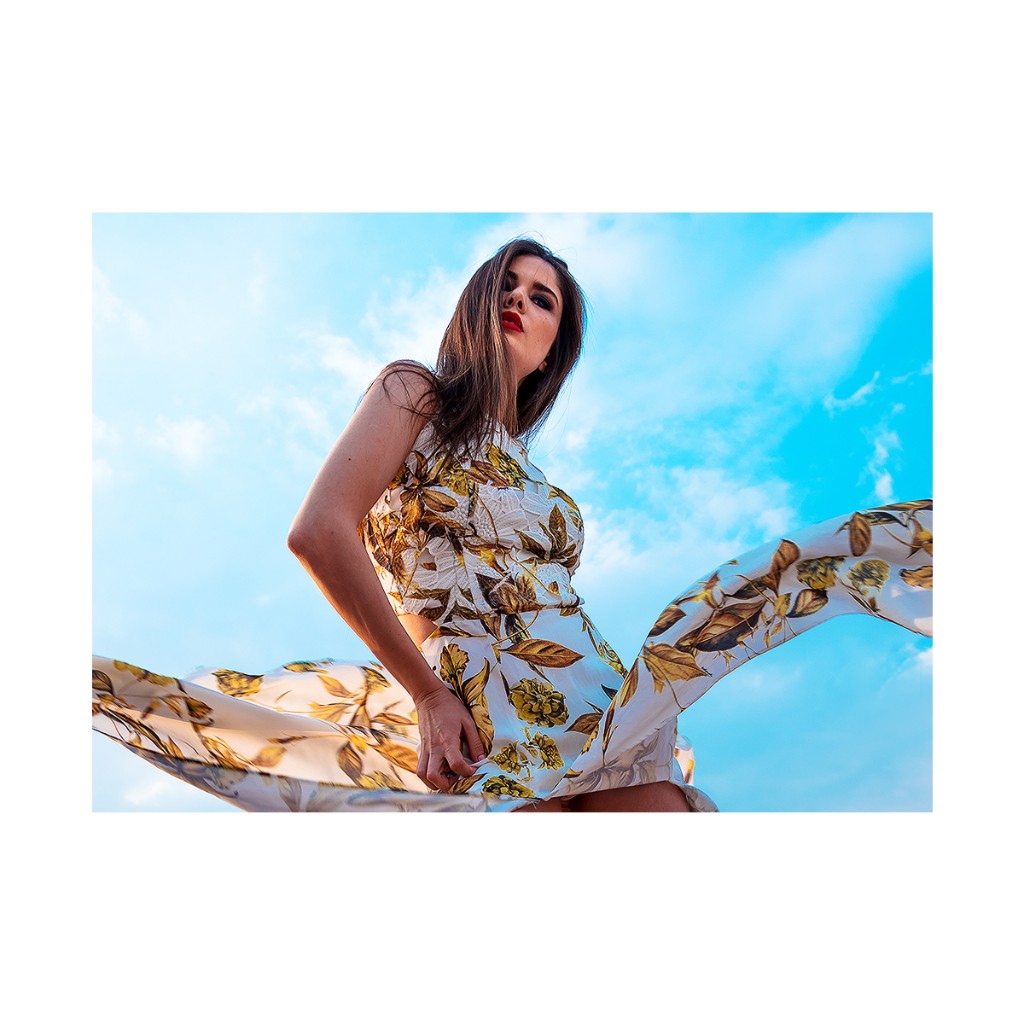 Ferezing editorial: floral dress with wind-blown wide skirt, looking down at camera, dramatic blue sky.