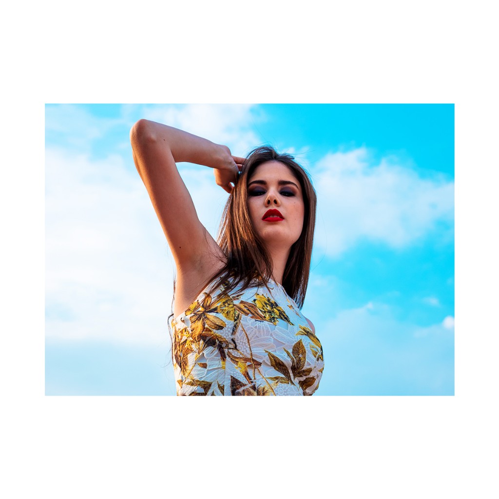 Ferezing portrait: low angle with white frame, bold red lip, yellow floral top, fine clouds in blue sky.