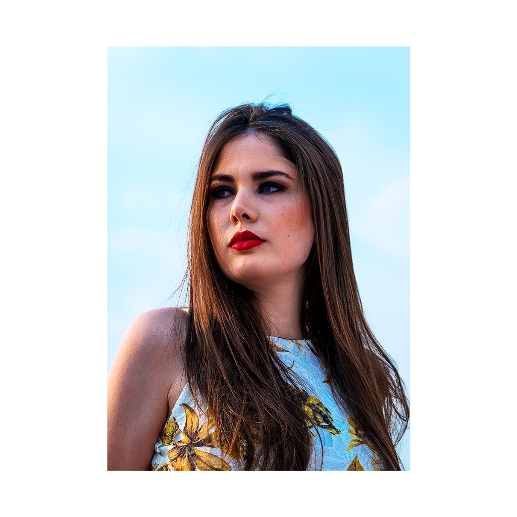 Ferezing close-up: gaze off-camera, matte red lip, gold floral on white, clean light-blue backdrop.