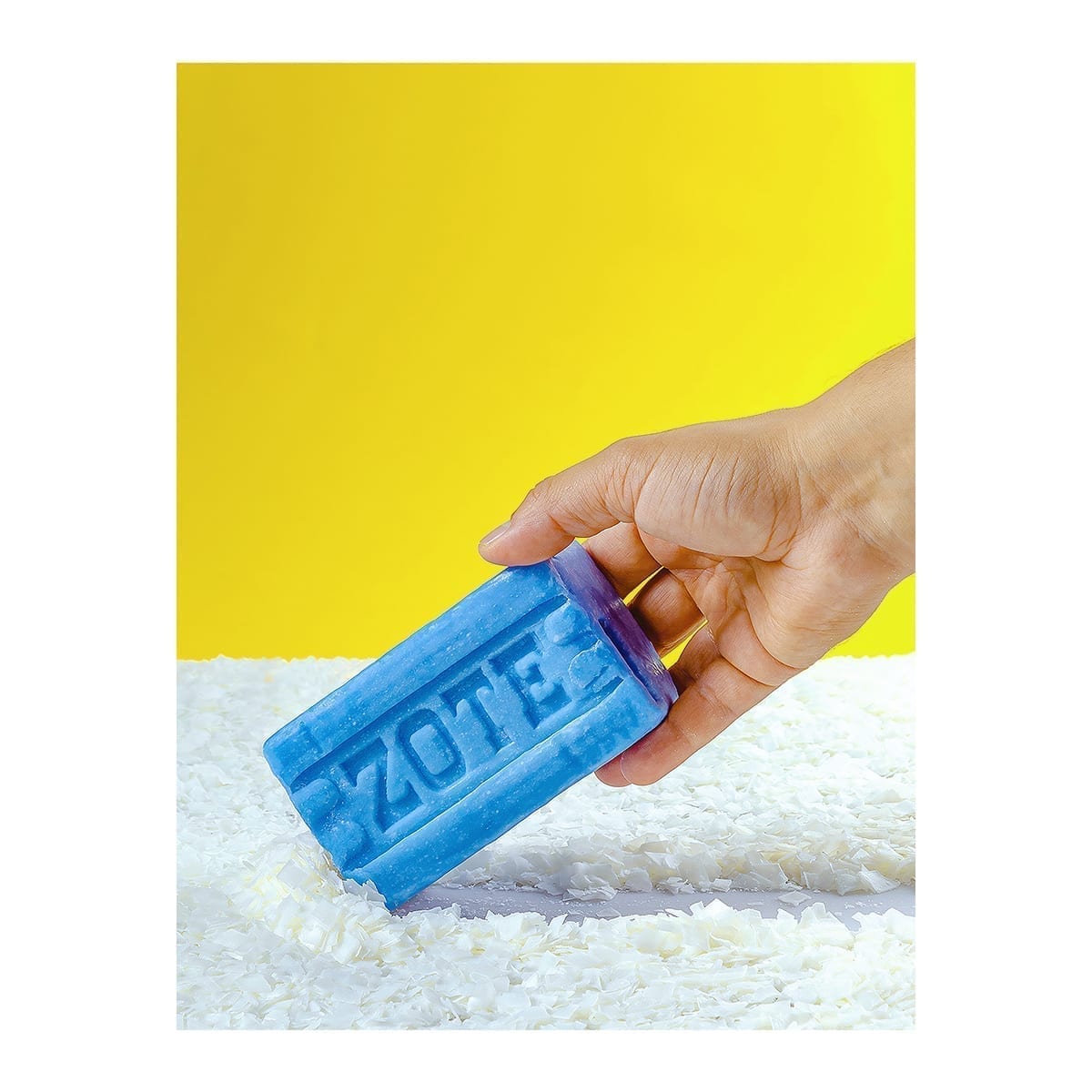 Blue Zote bar: hand pressing the bar through white shavings on yellow. “100 Years of History” editorial project, Fábrica de Jabón La Corona — piece 13 of 38.