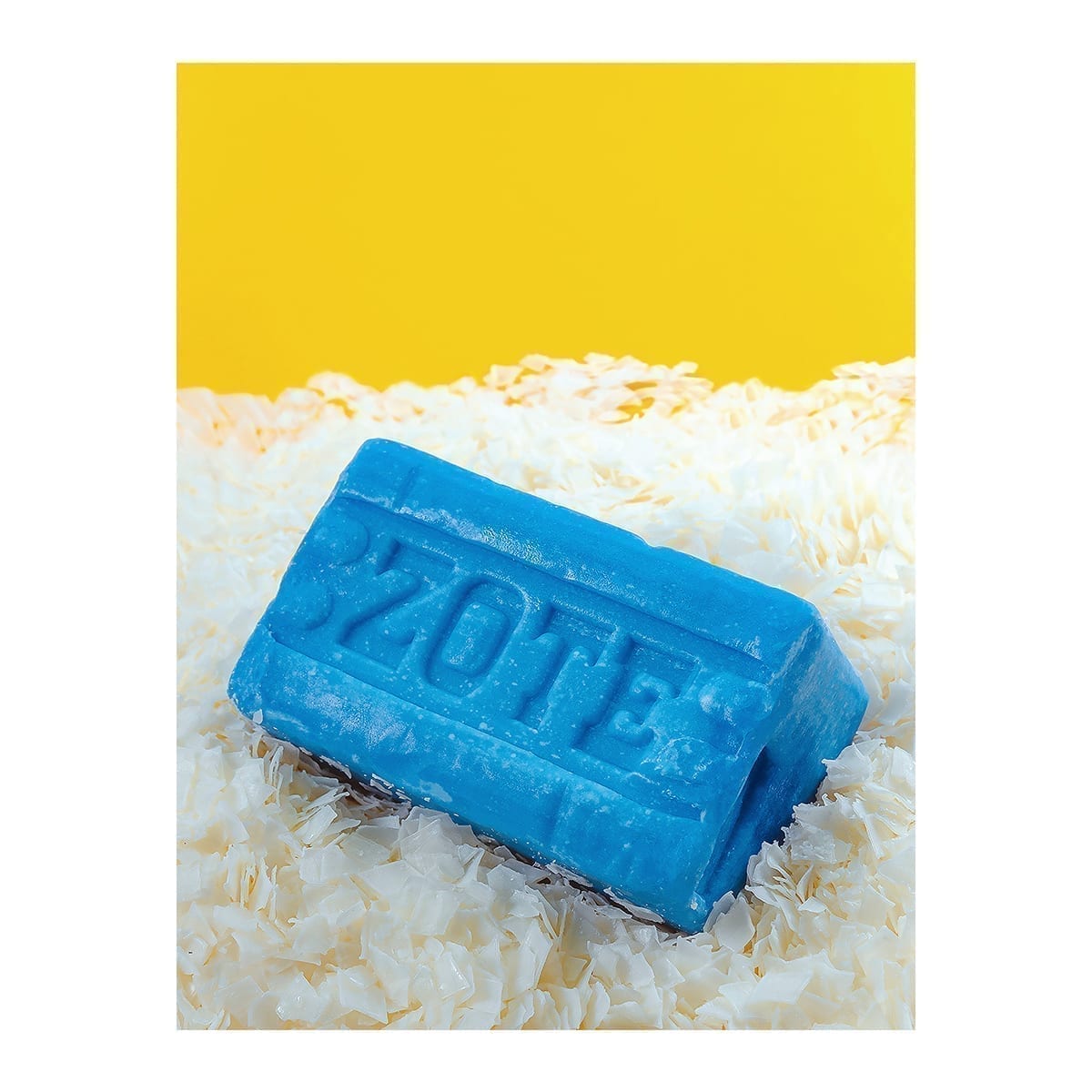 Blue Zote laundry bar on the diagonal over white shavings; flat yellow background. “100 Years of History” editorial project, Fábrica de Jabón La Corona — piece 14 of 38.