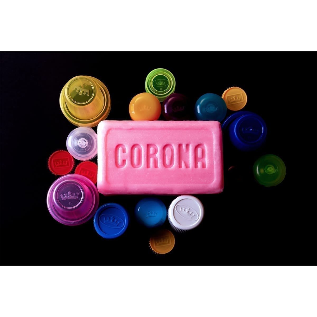 Pink soap bar embossed “CORONA” surrounded by multicolor plastic caps on a black backdrop. “100 Years of History” editorial project, Fábrica de Jabón La Corona — piece 2 of 38.