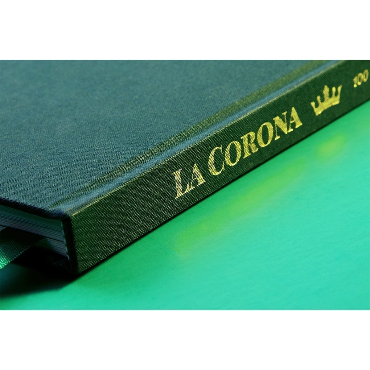 Book spine: LA CORONA, crown icon, and “100” in gold on green cloth; ribbon bookmark. “100 Years of History” editorial project, Fábrica de Jabón La Corona — piece 27 of 38.