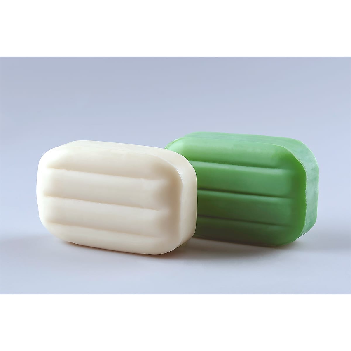 Two toilet soap bars (white and green) with horizontal grooves on a pale gray minimal set. “100 Years of History” editorial project, Fábrica de Jabón La Corona — piece 5 of 38.