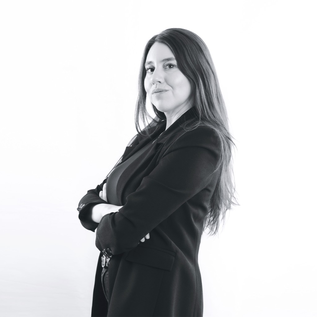 Liz Stryke B&W portrait: structured black blazer, arms crossed, three-quarter profile looking at camera; seamless white backdrop and soft light — corporate talent headshot.